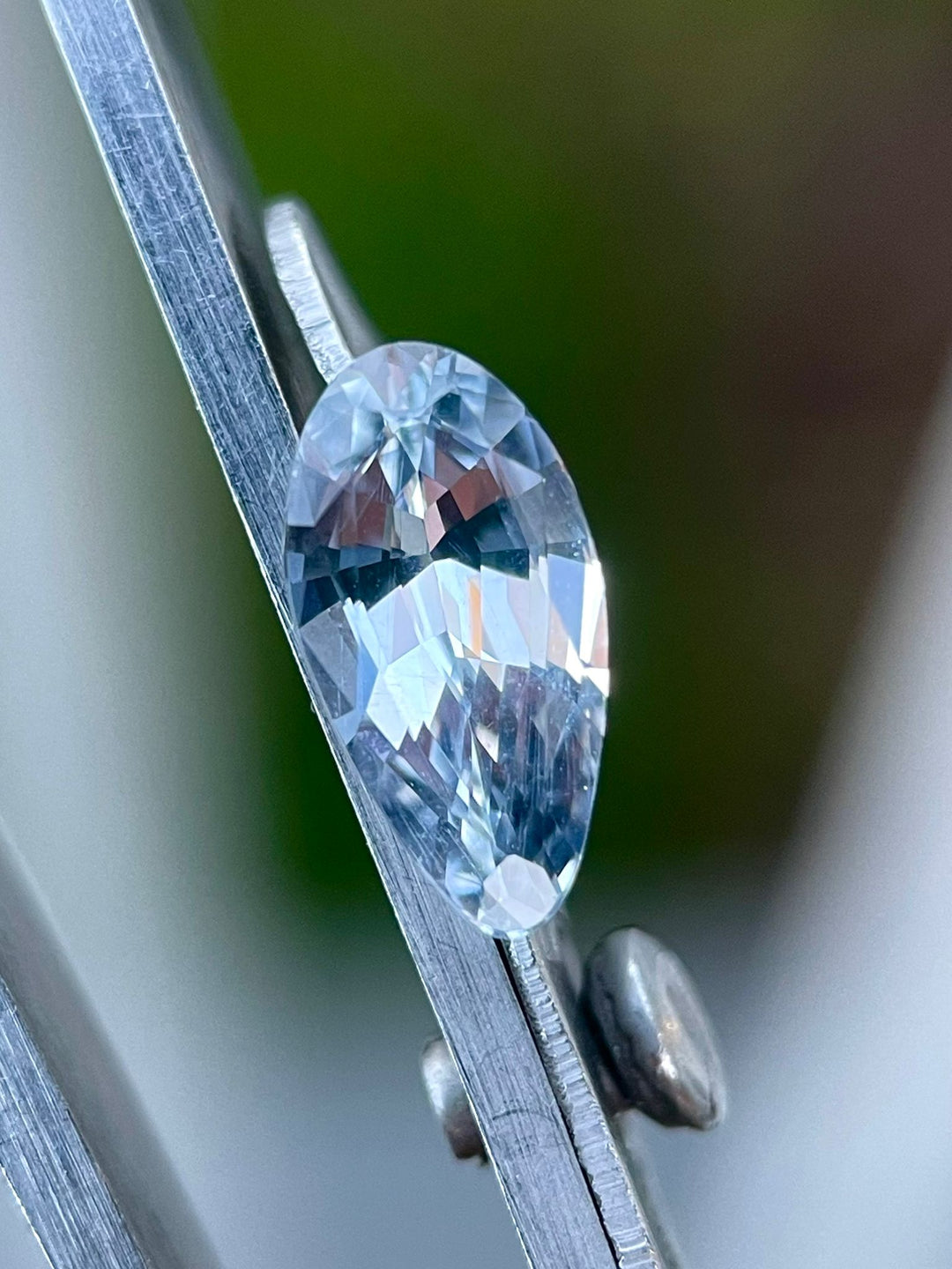 Detailed view of a light sage green Sri Lankan sapphire in tweezers, highlighting the precision of its custom modified pear shape and flawless cut. This 1.42 carat gemstone is showcased without any heat treatment, capturing its natural beauty and clarity.