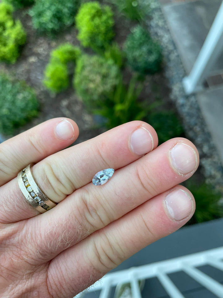 Outdoor shot of a hand holding a light sage green sapphire weighing 1.42 carats. The custom modified pear-shaped gemstone, sourced from Sri Lanka and untreated for color, sparkles against a blurred garden background.