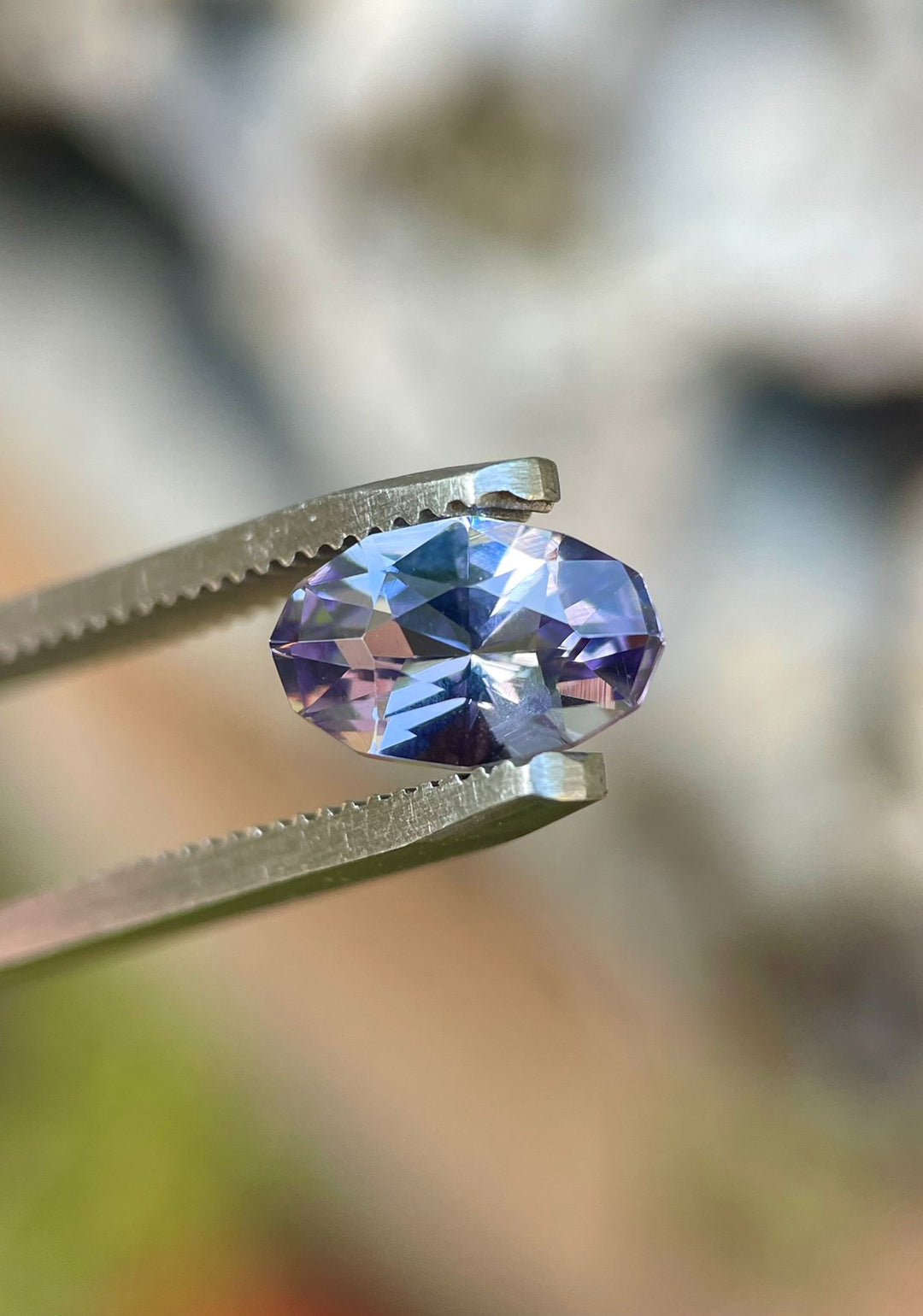 Close-up of a stunning 1.79 carat Tanzanite gemstone held in tweezers, showcasing its unique custom geometric oval shape and captivating gray, lavender, and purple hues. This premium Tanzanite is untreated and sourced from Merelani Hills, Tanzania.