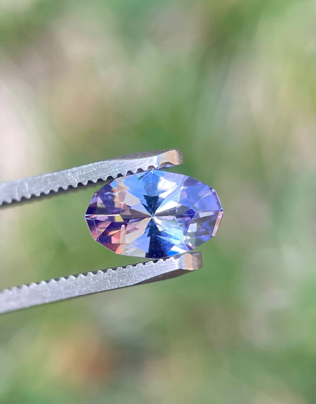 Detailed view of a 1.79 carat Tanzanite gemstone against a natural blurry green background, highlighting its custom geometric oval shape and a mix of gray, lavender, and purple colors. The gemstone's origin from Merelani Hills, Tanzania, enhances its rarity and value