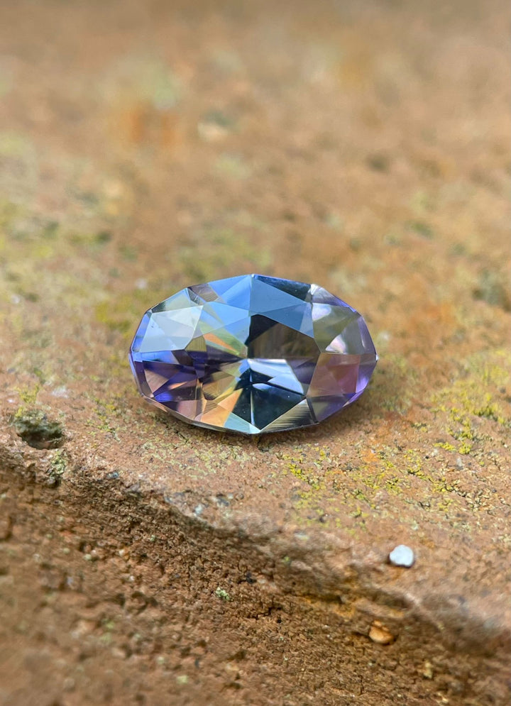 A 1.79 carat Tanzanite gemstone rests on an earthy surface, emphasizing its exquisite custom geometric oval shape and vibrant gray, lavender, and purple shades. Originating from the renowned Merelani Hills in Tanzania, this gemstone is presented in its natural, untreated state.