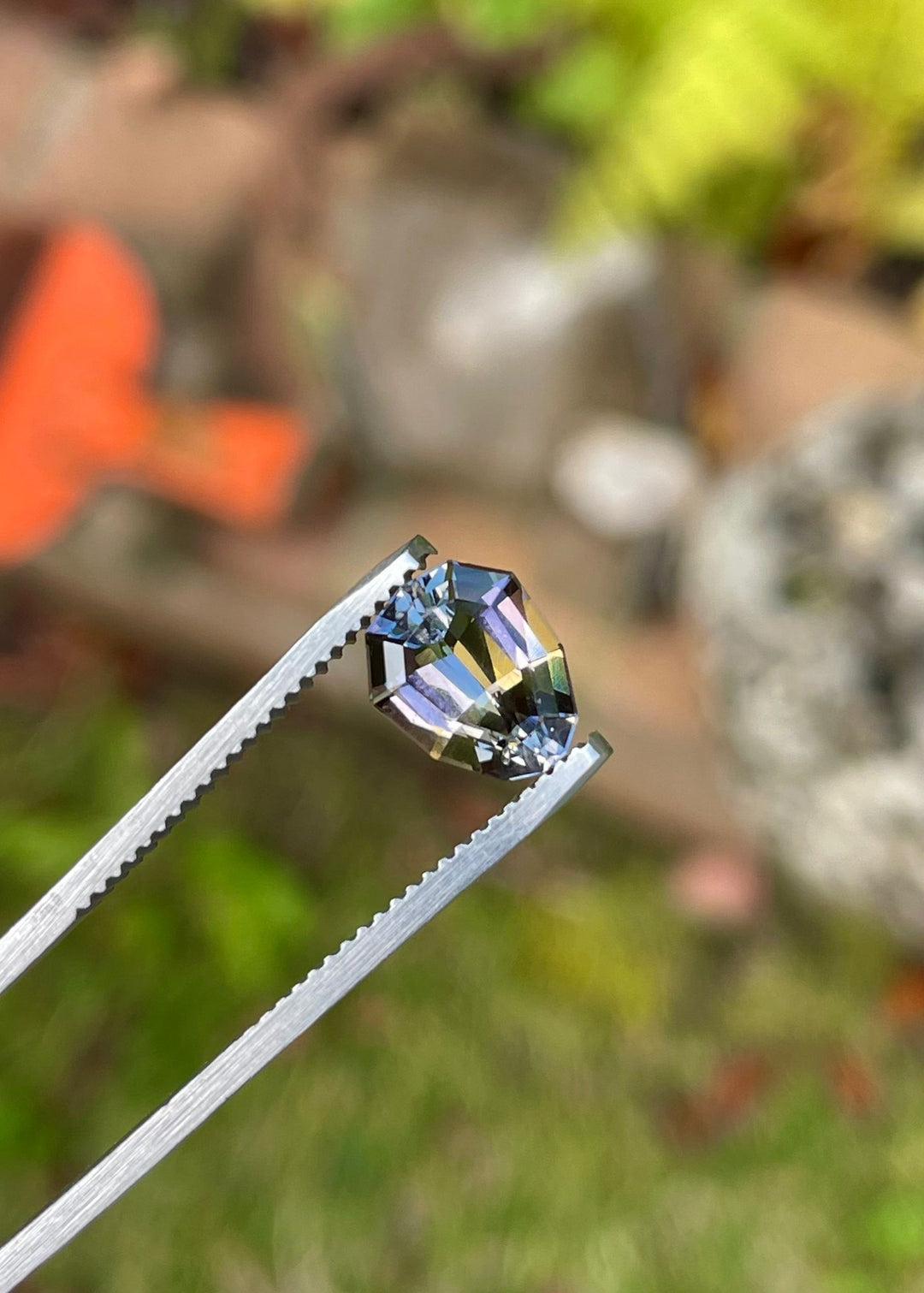 Close-up of a 2.10 carat Tanzanite held in tweezers against a blurred natural backdrop, highlighting its custom geometric pear shape and the colorful interplay of purple, yellow, and green. This Tanzanite originates from Merelani Hills, Tanzania, and remains untreated to preserve its natural beauty.