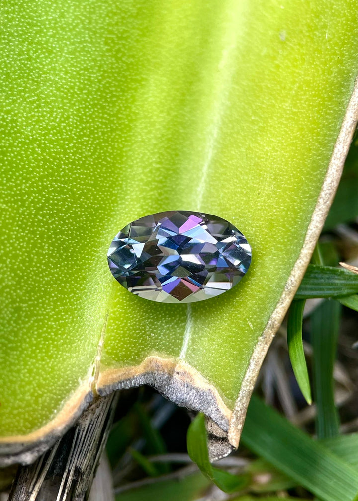 Close-up of a 1.50 carat Tanzanite resting on a green leaf, displaying its exceptional green, teal, and purple shades. The custom oval cut and the natural backdrop emphasize the gemstone's origin from Merelani Hills, Tanzania, and its natural beauty.