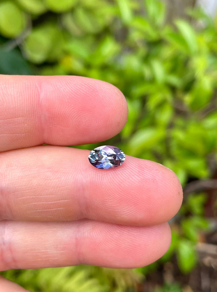 A stunning 1.50 carat Tanzanite gemstone held between fingers, showcasing a vibrant mix of green, teal, and purple colors. This custom mix cut oval Tanzanite, untreated and from Merelani Hills, Tanzania, highlights its unique multicolor hue and exquisite cut.
