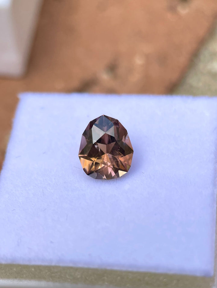 A 2.43 carat Tanzanite gemstone in a custom geometric pear shape displayed on a blue background, showcasing rich brown and champagne colors. Originating from Merelani Hills, Tanzania, this untreated gemstone exemplifies unique craftsmanship and natural beauty.