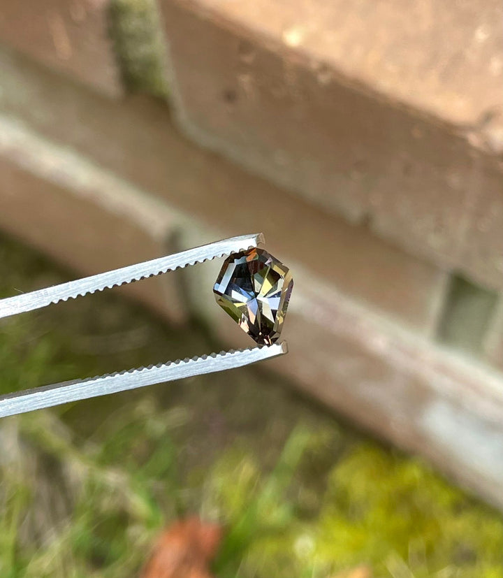 Close-up view of a 1.41 carat Tanzanite gemstone held in tweezers against a rustic outdoor background, emphasizing its intricate geometric mix cut shield shape and captivating color blend of yellow, green, red-orange, and brown. This Tanzanite is a natural gem from Tanzania, showcasing exceptional craftsmanship.