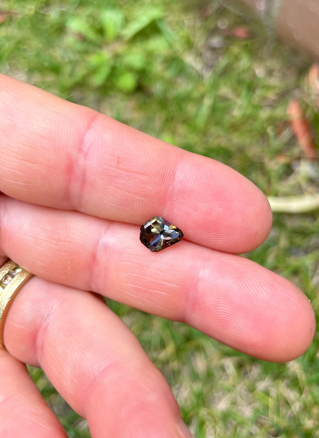 Hand-held 1.41 carat Tanzanite gemstone, featuring a stunning array of yellow, green, red-orange, and brown colors in a custom geometric mix cut shield shape. This exquisite gem from Merelani Hills, Tanzania, is displayed against a natural outdoor backdrop, accentuating its rich color palette and precise cut.