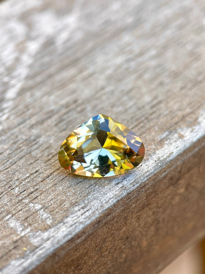Close-up of a Tanzanite gemstone resting on a rustic surface, highlighting its unique teal, gold, and yellow colors. This 2.48-carat Tanzanite, with dimensions of 8.07 x 11.16 x 4.79 mm, boasts a wide pear brilliant cut, originating from the Arusha Region in Tanzania, priced at $818.40.