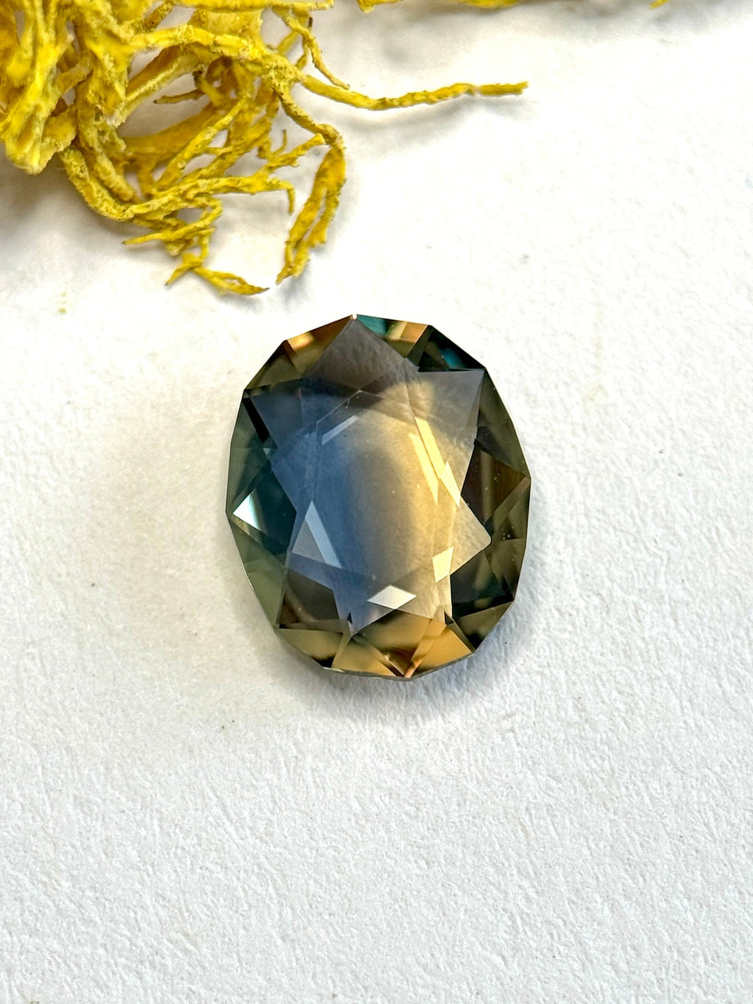 Oval portrait cut parti sapphire on white background, revealing strong angular facet structure and distinct blue-to-yellow color zoning with reflective flashes along the edges.