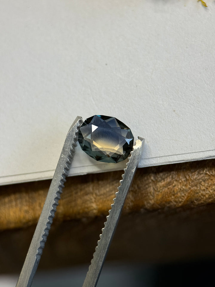 1.13ct blue and yellow parti sapphire held in silver tweezers, showing a sharp division of dark teal-blue across the crown and warm golden-yellow reflection near the center, with crisp geometric portrait facets.