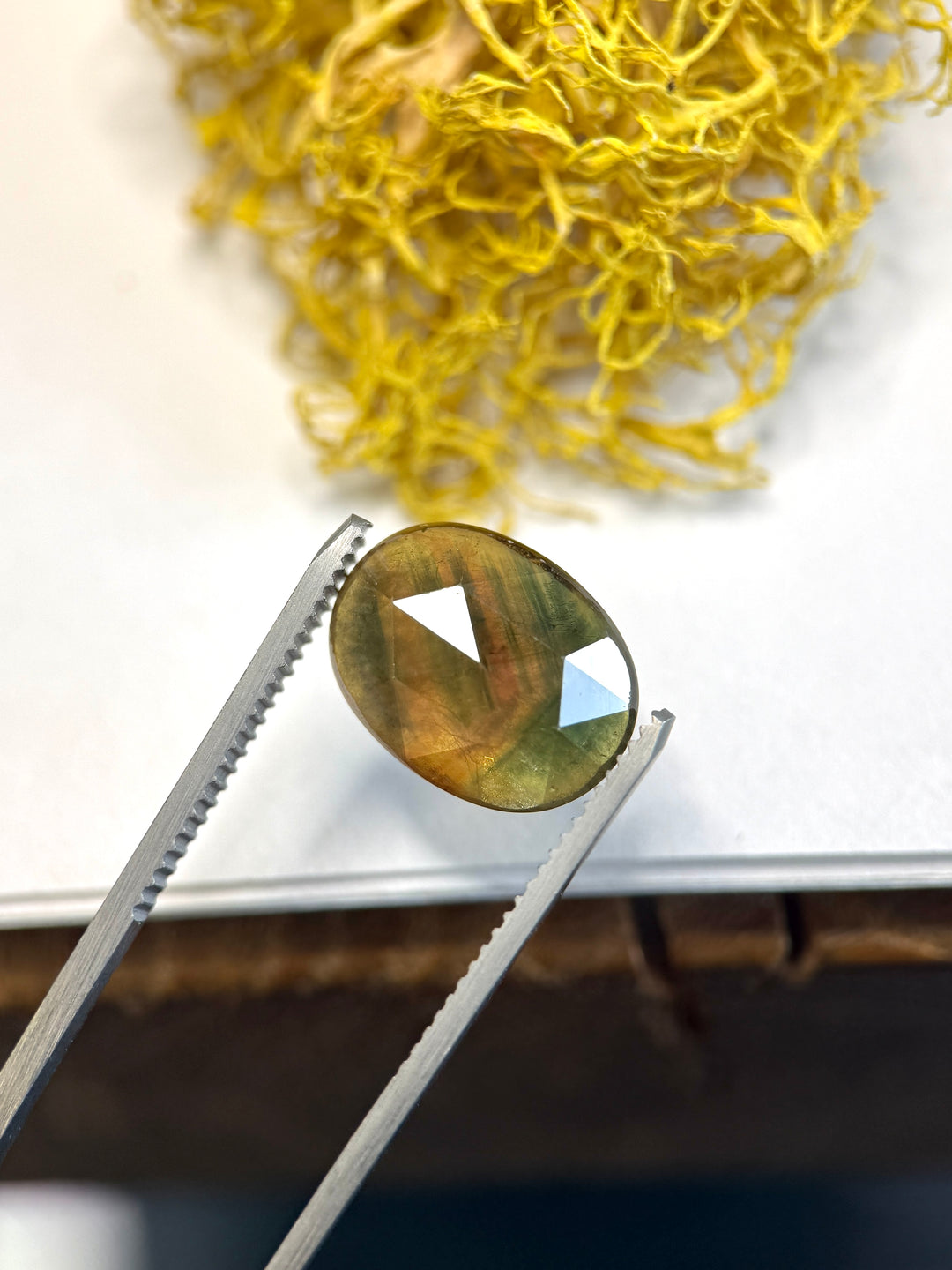 8.17ct orange and green parti sapphire held in metal tweezers, displaying broad rose-cut facets and visible natural internal silk with earthy moss and amber tones.