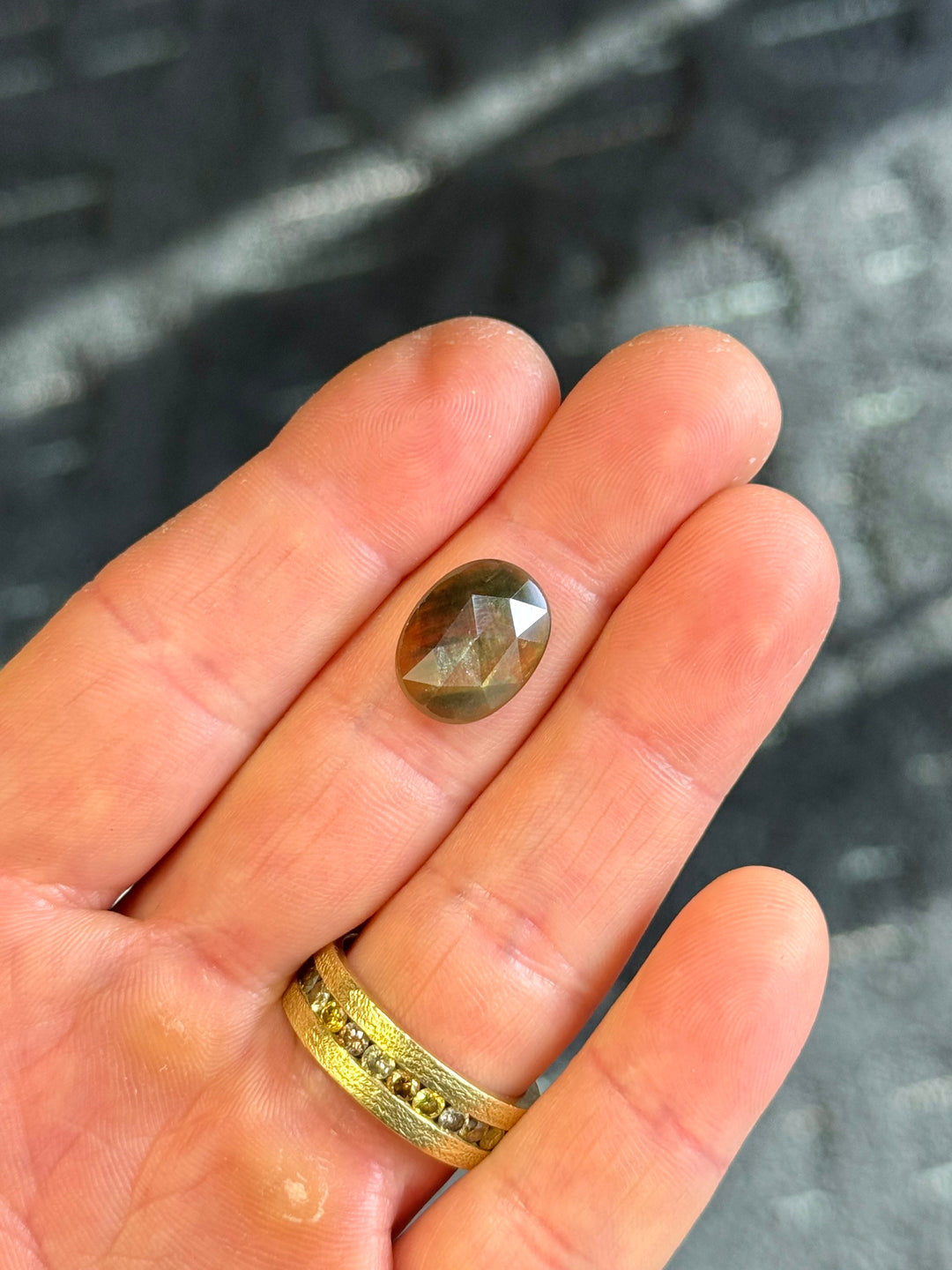 Oval rose-cut Thai sapphire on fingertip, revealing painterly streaks of green and burnt orange with soft surface reflections and subtle inclusions.