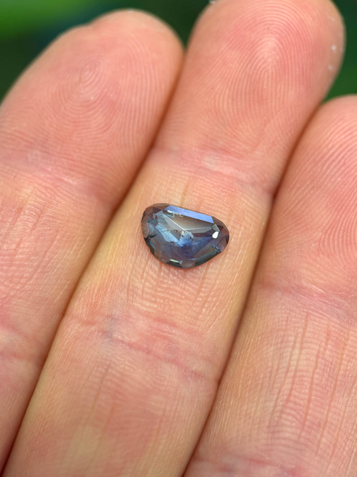 1.73ct blue and yellow parti sapphire resting on fingertip, showing deep navy-blue body color with soft golden-yellow zoning across a custom triangular rose-cut surface.