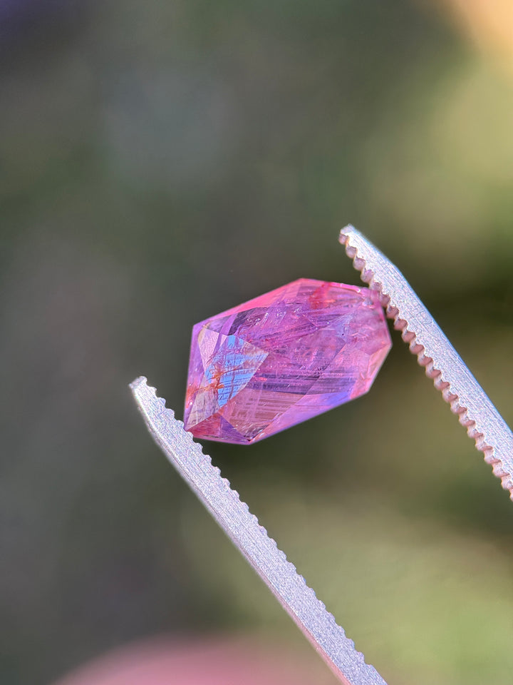Elongated free-form pink and purple sapphire held in metal tweezers, showing vivid magenta body color with visible internal silk and angular asymmetrical facets.