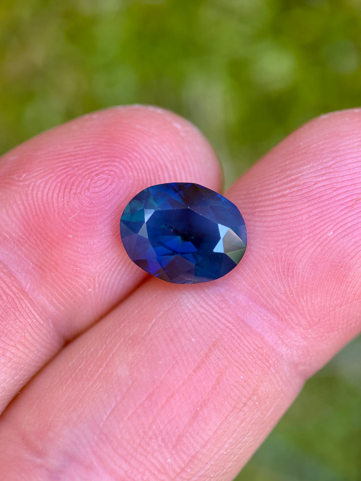 4.60ct deep royal teal blue sapphire resting on fingertips, showing saturated blue-green color and visible internal silk under natural light.