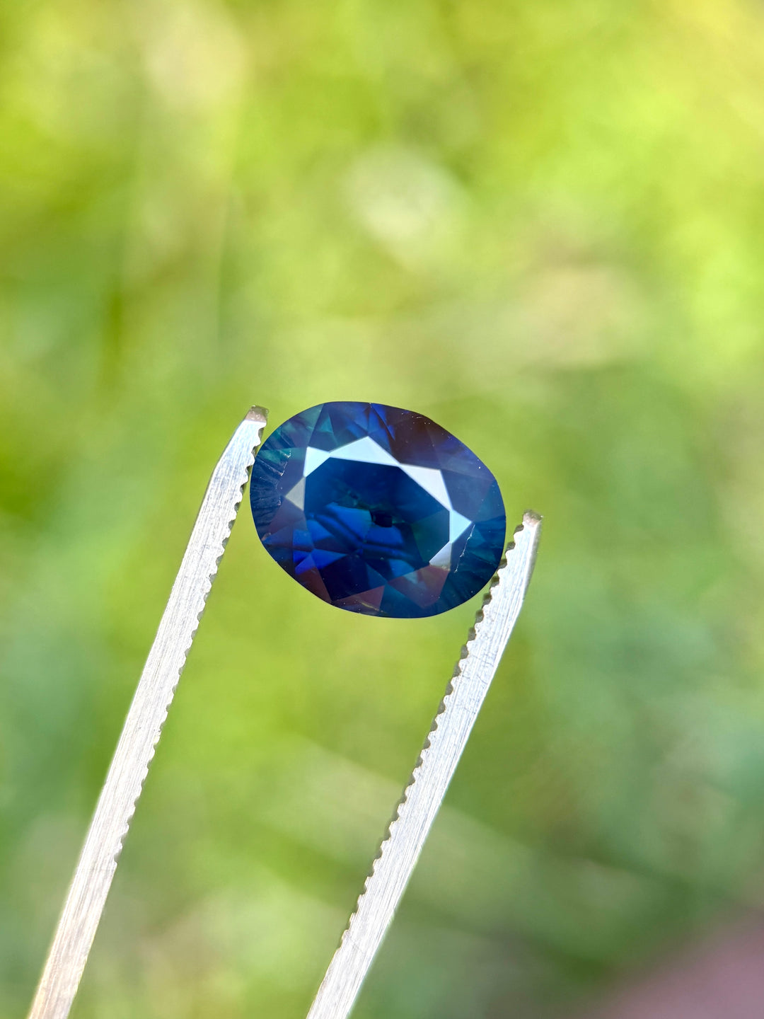 Unheated Nigerian sapphire held in metal tweezers, displaying rich inky teal-blue tones with brilliant facet reflections.
