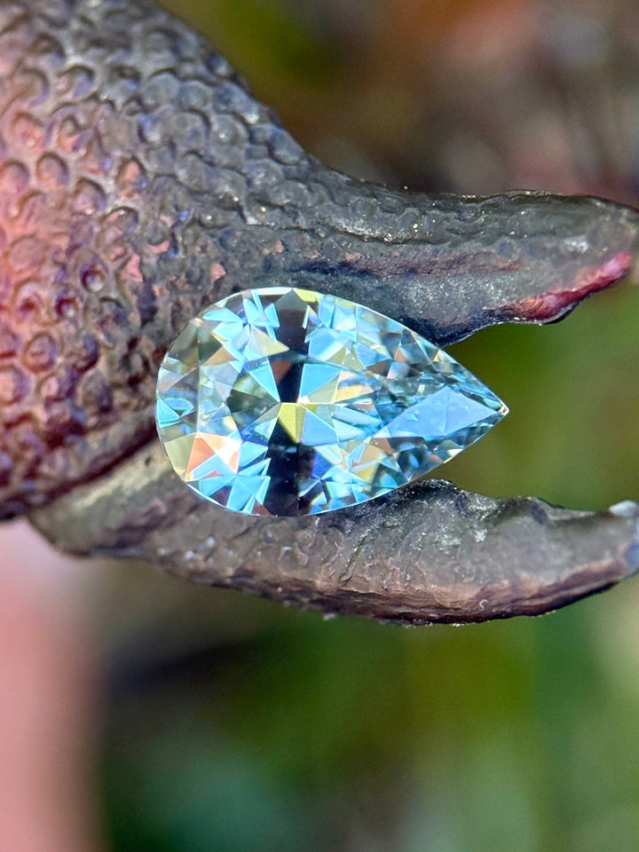 Precision pear periwinkle sapphire nestled against textured natural surface, displaying pastel blue-violet tones and brilliant internal sparkle.