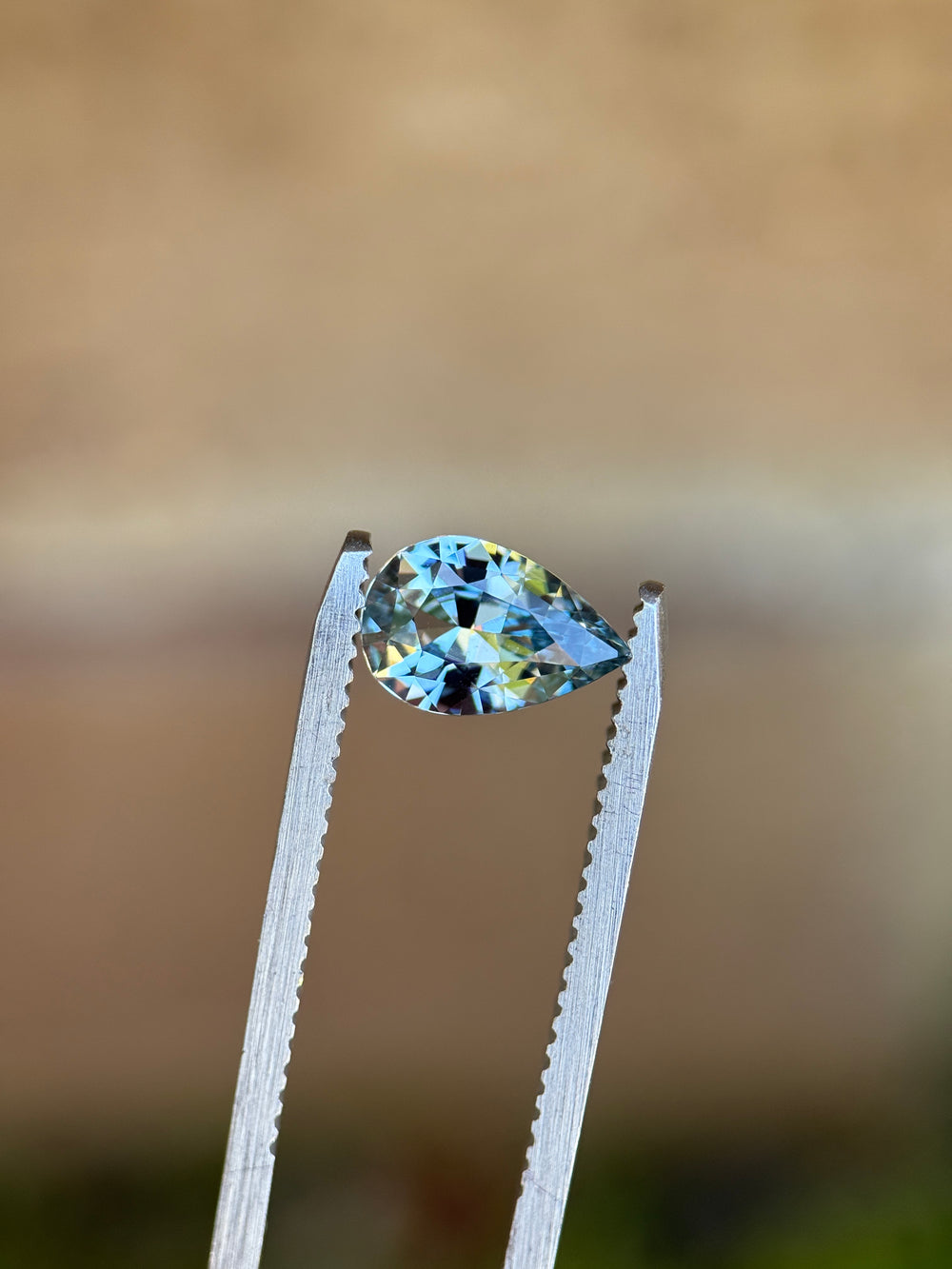 Unheated periwinkle pear sapphire held in silver tweezers against a neutral background, revealing sharp facet junctions and lively blue and lavender reflections.