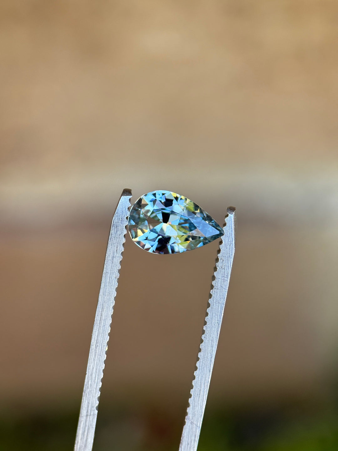 Unheated periwinkle pear sapphire held in silver tweezers against a neutral background, revealing sharp facet junctions and lively blue and lavender reflections.