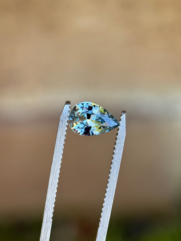 Unheated periwinkle pear sapphire held in silver tweezers against a neutral background, revealing sharp facet junctions and lively blue and lavender reflections.