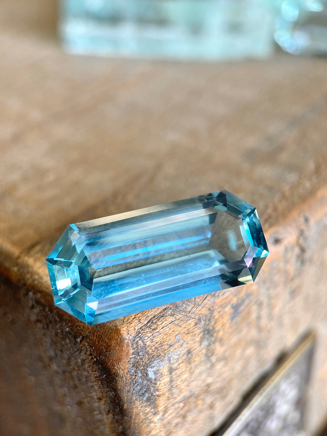 Stunning 11.66-carat aquamarine gemstone showcased on a rustic wooden surface, emphasizing its double blue color and elongated emerald cut. This high-quality aquamarine, sourced from Nasarawa, Nigeria, displays its impressive dimensions and natural, untreated clarity, highlighted by the play of light across its precise facets.