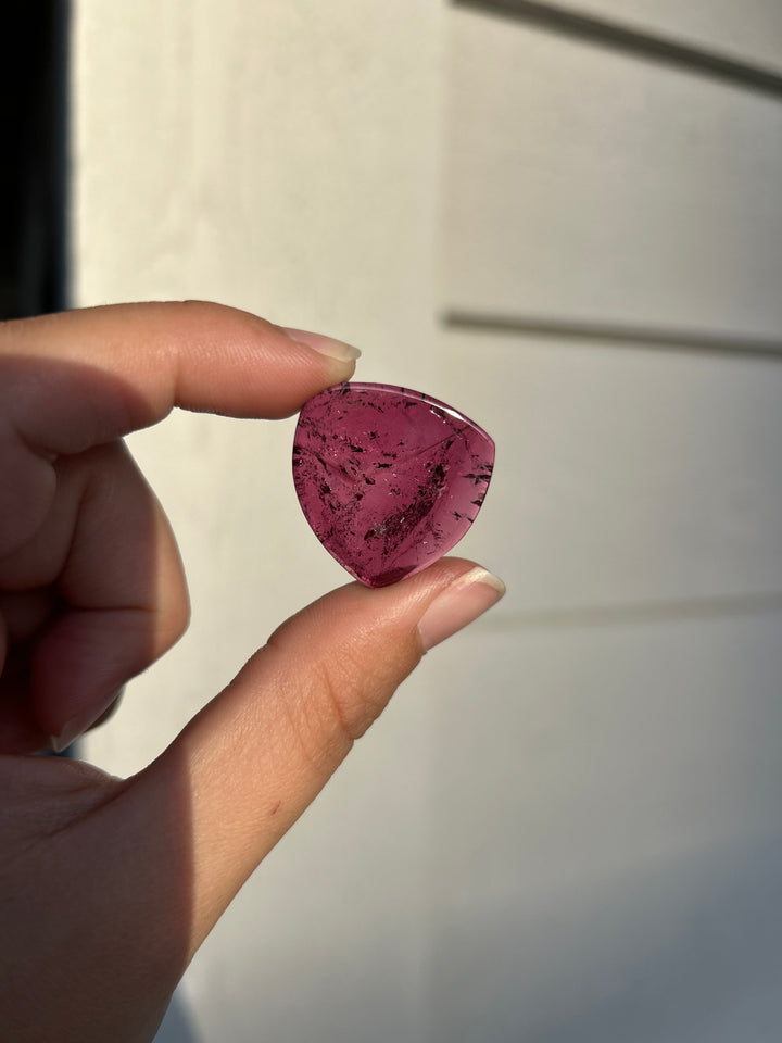A 28.02 carat natural deep pink tourmaline slice from Congo, held between fingertips in direct sunlight. Its translucent raspberry-pink crystal reveals dark natural inclusions and intricate internal textures within a softly triangular polished silhouette.