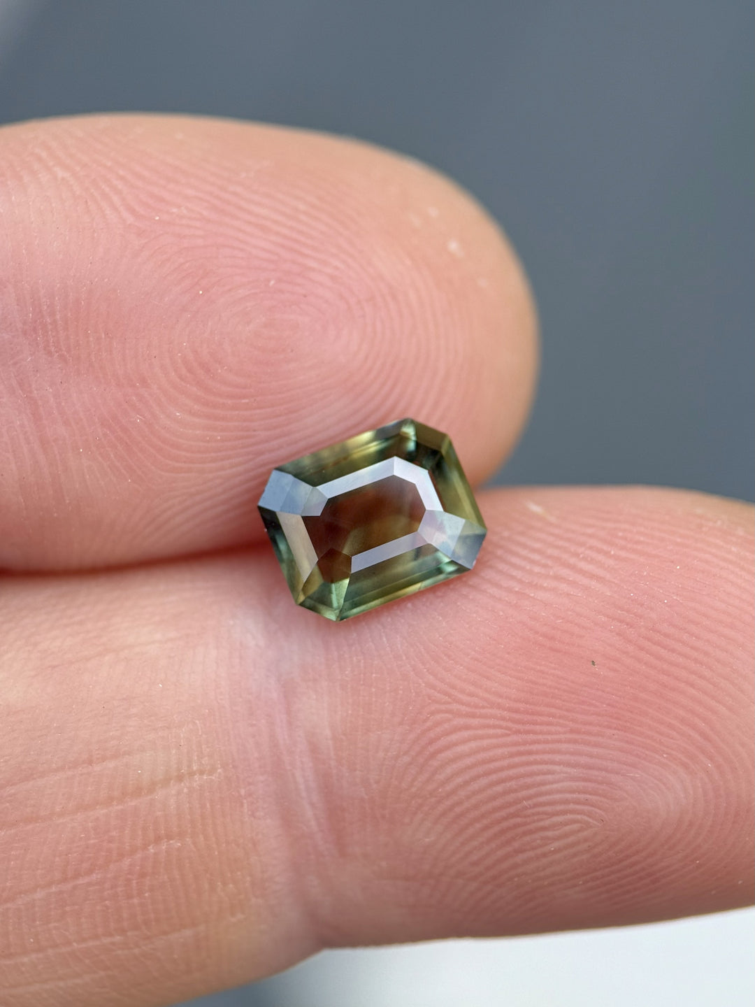 Natural Kenyan sapphire in emerald cut, viewed on fingertips, showcasing vibrant interplay of brown, gold, and forest green hues – 2.02 carats.