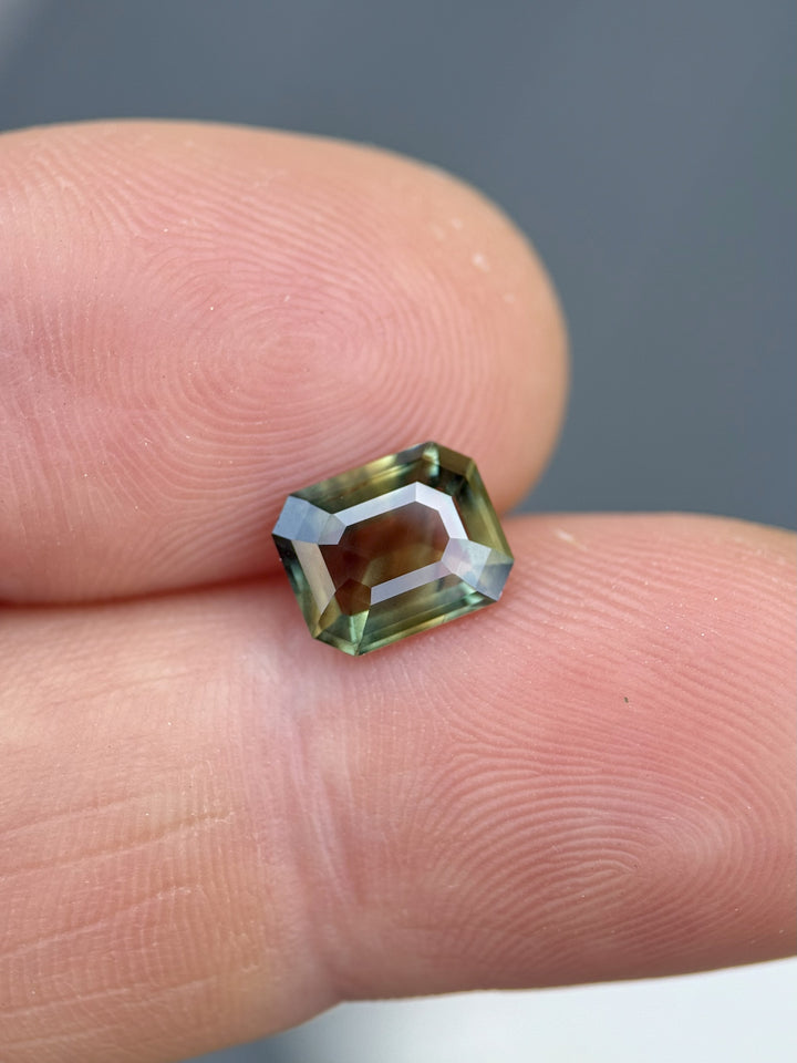 Natural Kenyan sapphire in emerald cut, viewed on fingertips, showcasing vibrant interplay of brown, gold, and forest green hues – 2.02 carats.
