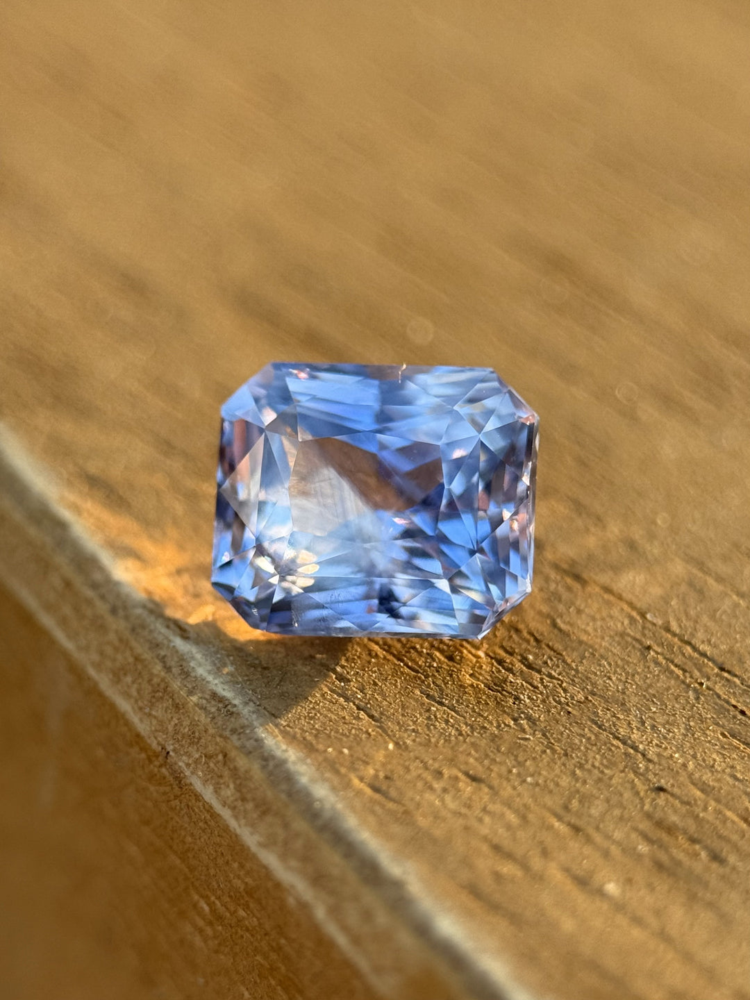 Close-up of a 2.70-carat custom radiant cut sapphire with lavender, violet, and blue tones, shown on a wooden surface.