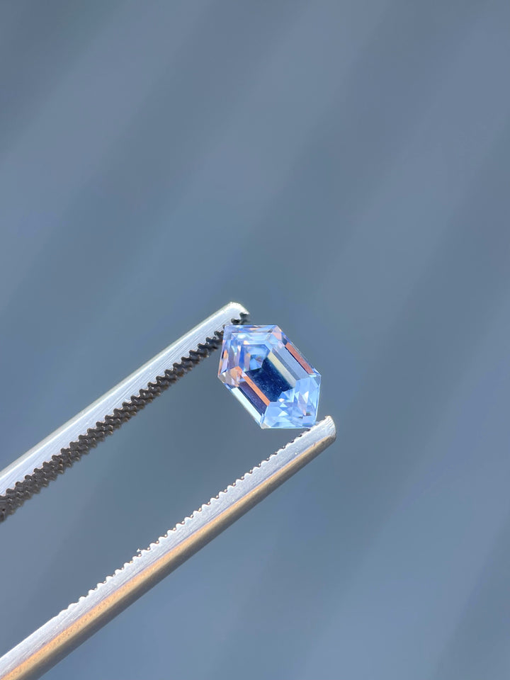 A 1.62-carat light Ceylon blue sapphire is held by precision tweezers. The step-cut hexagonal gemstone glistens under natural light, showing its clean lines and sharp facets. This untreated, no-heat sapphire measures 8.78mm by 5.06mm by 4.50mm and is sourced from Sri Lanka.