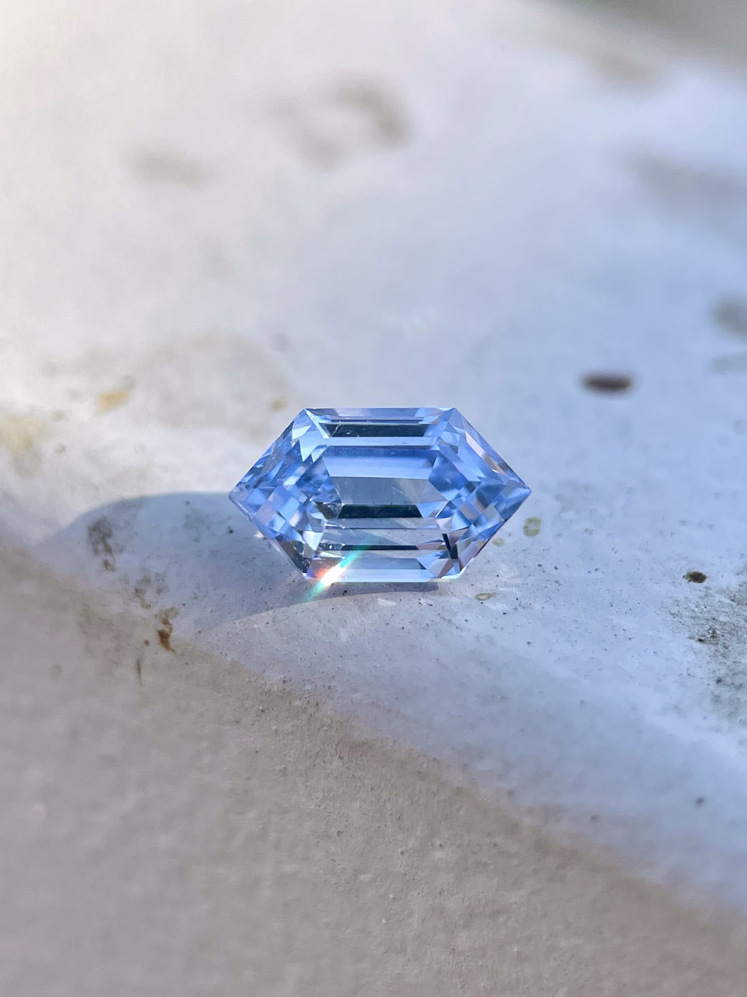A 1.62-carat light Ceylon blue sapphire sits elegantly on a flat stone surface, showcasing its step-cut hexagonal design. The gemstone's clean facets catch the light, revealing its clarity and untreated origin. The sapphire, from Sri Lanka, has dimensions of 8.78mm by 5.06mm by 4.50mm.