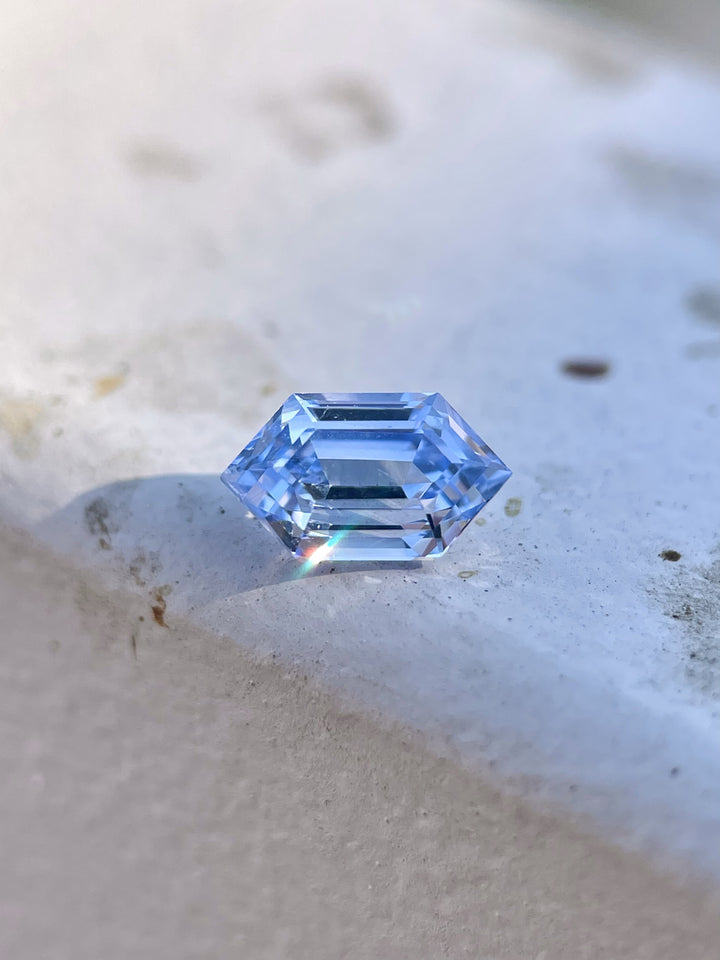A 1.62-carat light Ceylon blue sapphire sits elegantly on a flat stone surface, showcasing its step-cut hexagonal design. The gemstone's clean facets catch the light, revealing its clarity and untreated origin. The sapphire, from Sri Lanka, has dimensions of 8.78mm by 5.06mm by 4.50mm.