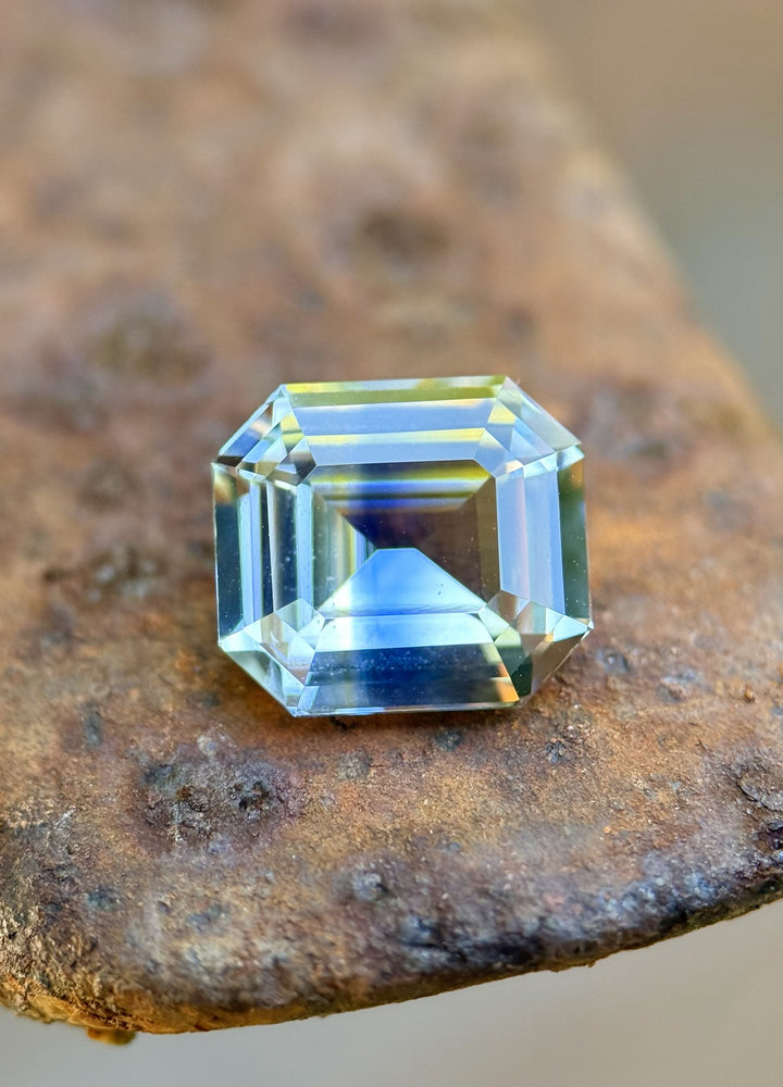 4.04 carat light yellow and blue parti sapphire in a precision emerald cut resting on textured metal, showcasing clean step facets and natural color zoning from Sakaraha, Madagascar.