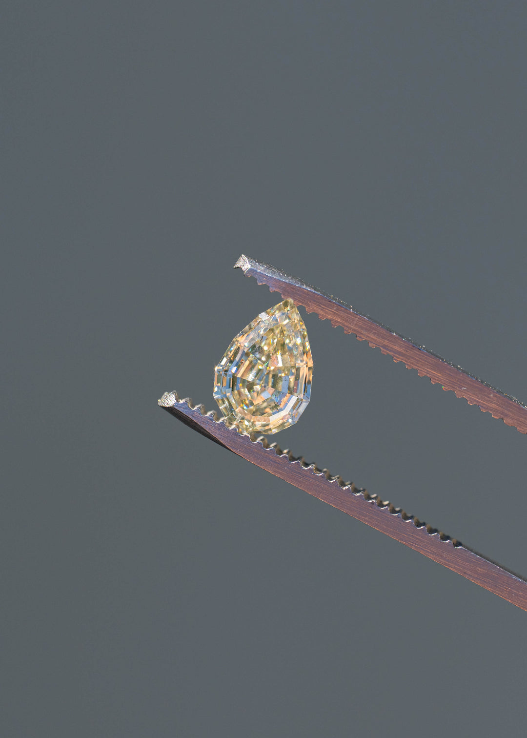 Light Yellow Geo Pear step cut diamond held in tweezers against a gray background, showcasing broad reflective facets, natural inclusions, and warm champagne-yellow brilliance.