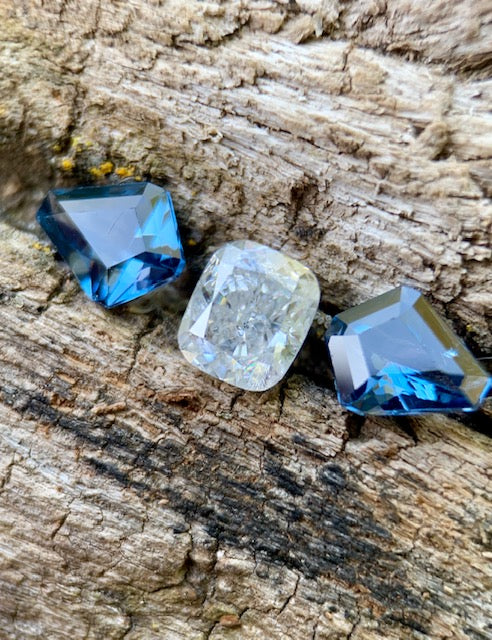 Three blue and white faceted loose gemstones on natural wood, showcasing brilliance and contrast for custom jewelry and designer sourcing.