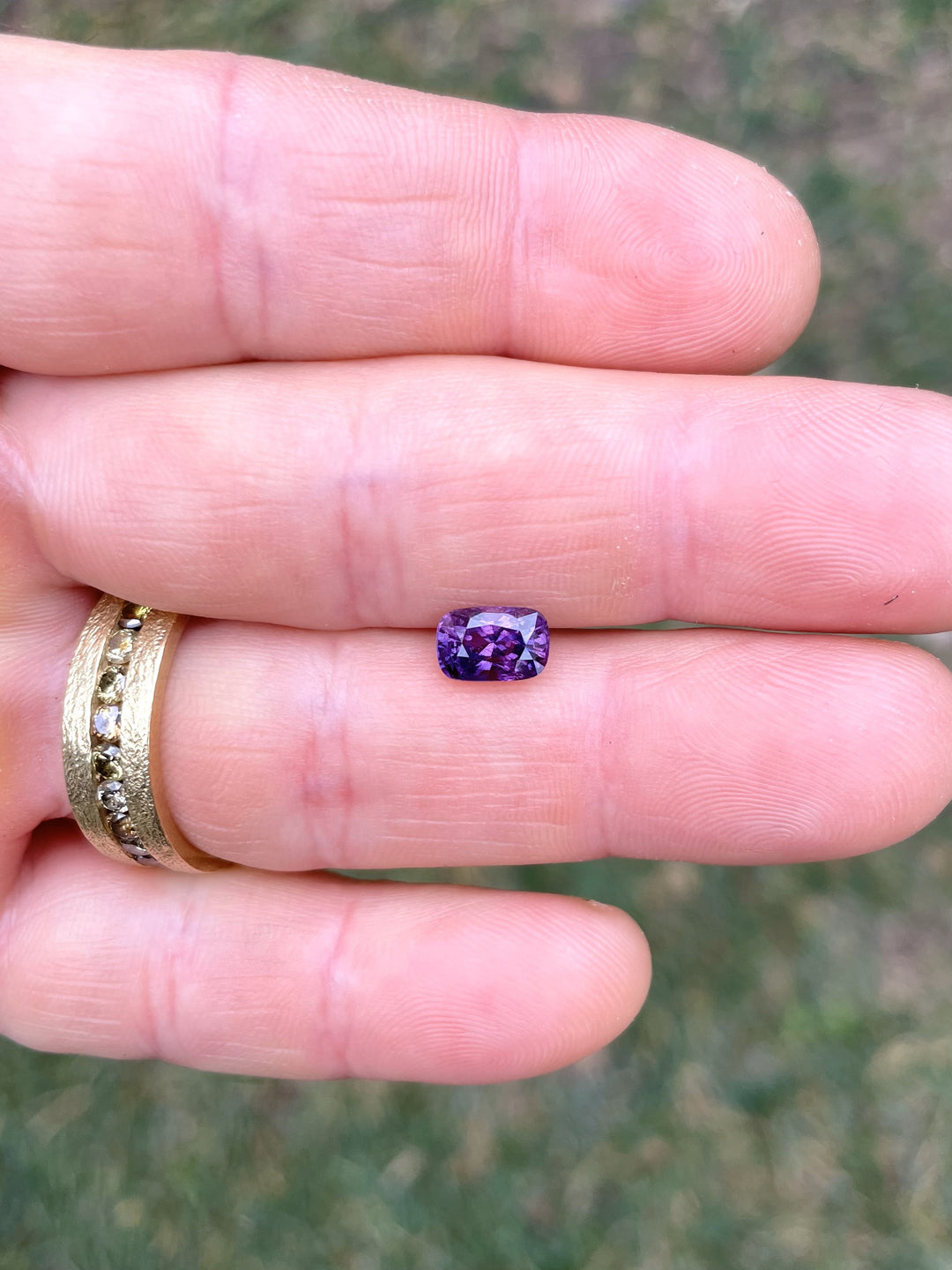 Close-up view of a 1.75-carat pink and mulberry sapphire with a mix cushion cut, shown on a person's fingertip against a grassy background. The stone's vibrant colors and fine cut bring out its exquisite, natural luster, emphasizing the untreated nature of this rare gemstone from Luc Yen, Vietnam.