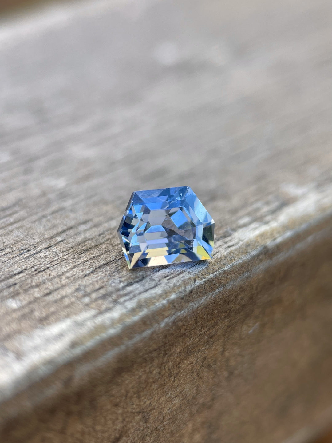 A 1.42 carat No Heat Sapphire with a custom geometric step cut, displayed on a rustic wooden surface. The sapphire exudes a deep cornflower blue color, boasting its unheated, pure state from Sri Lanka, illustrating both brilliance and depth