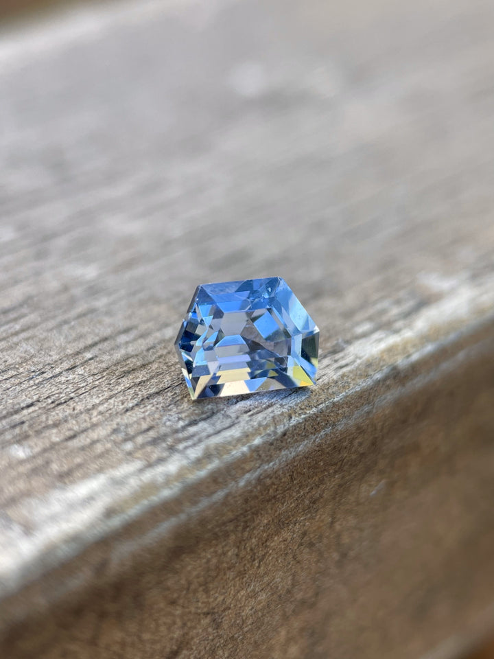 A 1.42 carat No Heat Sapphire with a custom geometric step cut, displayed on a rustic wooden surface. The sapphire exudes a deep cornflower blue color, boasting its unheated, pure state from Sri Lanka, illustrating both brilliance and depth