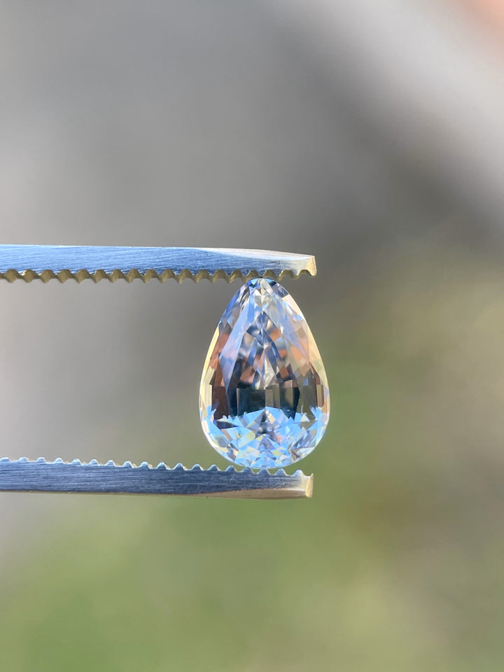 A breathtaking 2.39-carat cornflower blue sapphire held delicately in tweezers, captured in natural lighting that enhances its deep, vivid colors and brilliant teardrop shape.