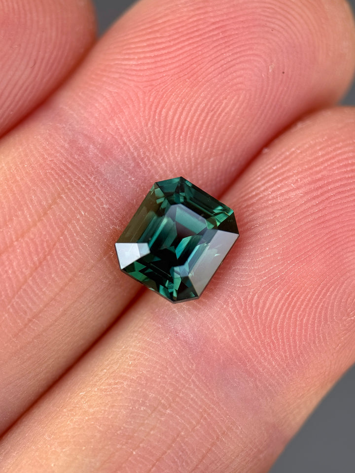 Loose vivid blue green sapphire resting on fingertips showing the emerald cut and color shift between blue and green tones.