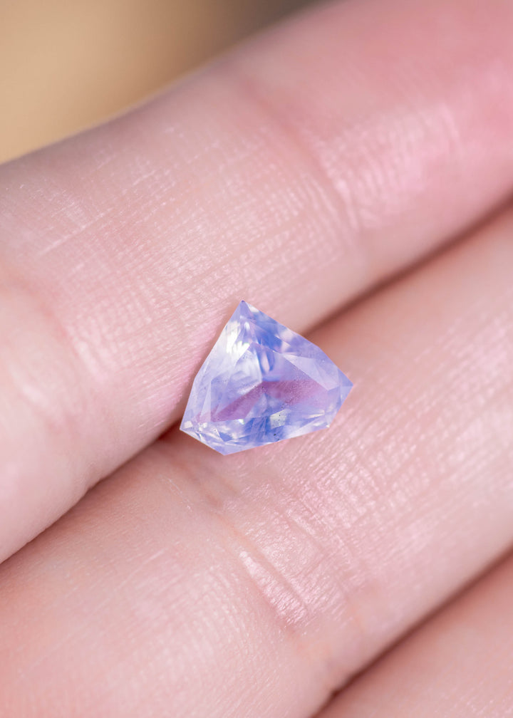 Loose opalescent blue sapphire resting on fingertips showing the custom geometric shield cut and subtle blue glow in natural light.