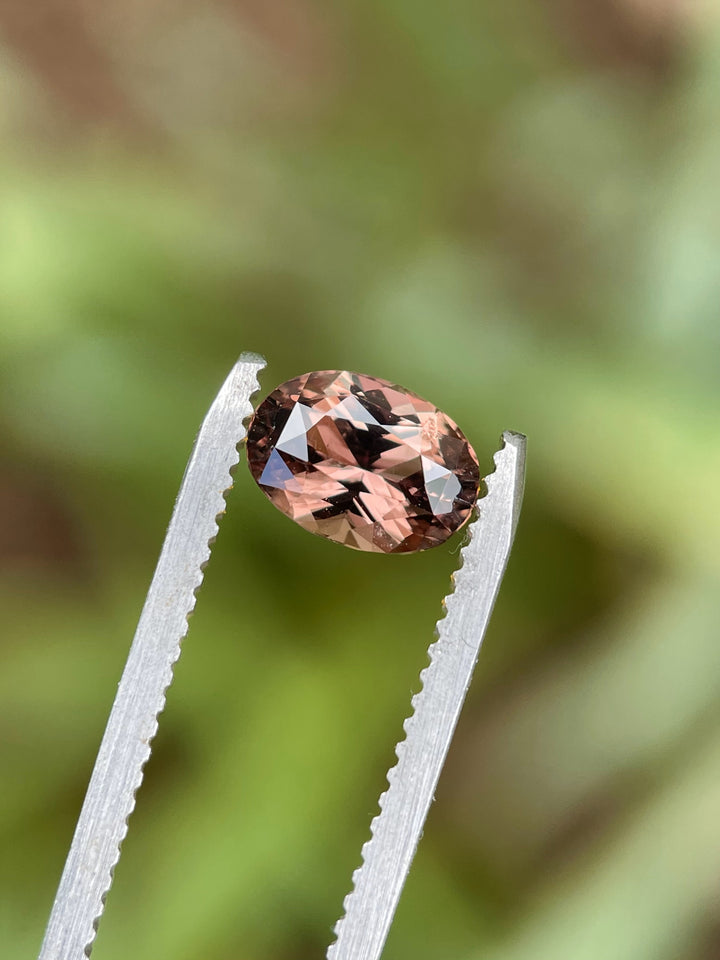 A 1.68-carat oval brilliant color-change sapphire held in gem tweezers, transitioning between purplish pink and brownish hues with a soft green backdrop.