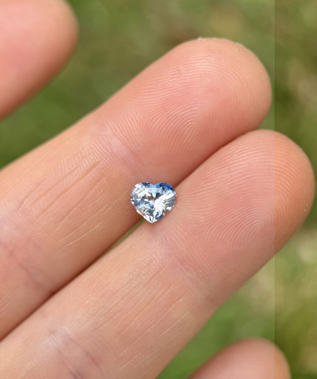 Natural sapphire in a brilliant heart shape, weighing 1.30 carats, displayed on fingertips with blue and colorless parti tones – heated gem from Madagascar.