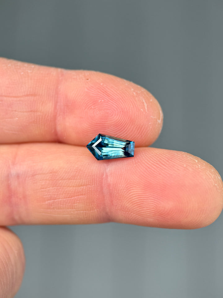 2.11-carat banded teal blue parti sapphire shown on a fingertip to demonstrate size and brilliance, featuring a precision elongated shield step cut from Madagascar.