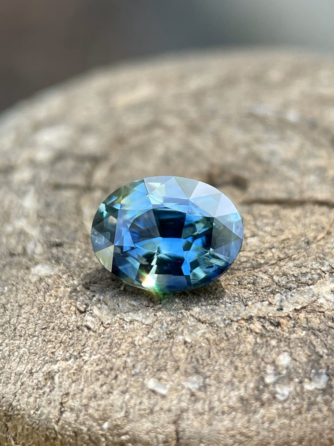 A vivid 1.30-carat natural sapphire with a mix of teal, green, and blue colors, showcased in a close-up on a rustic stone surface.
