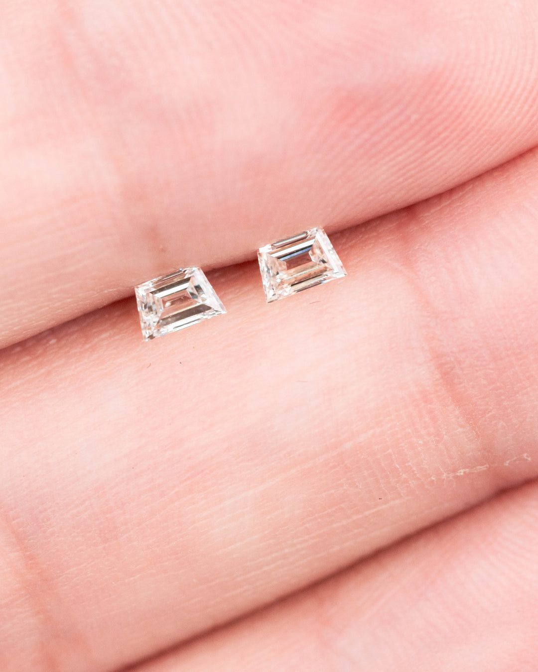 0.29 carat matched pair of G–H color VVS clarity trapezoid step cut diamonds displayed on fingertips, showing the clean step faceting, symmetry, and brilliance of untreated South African diamonds.