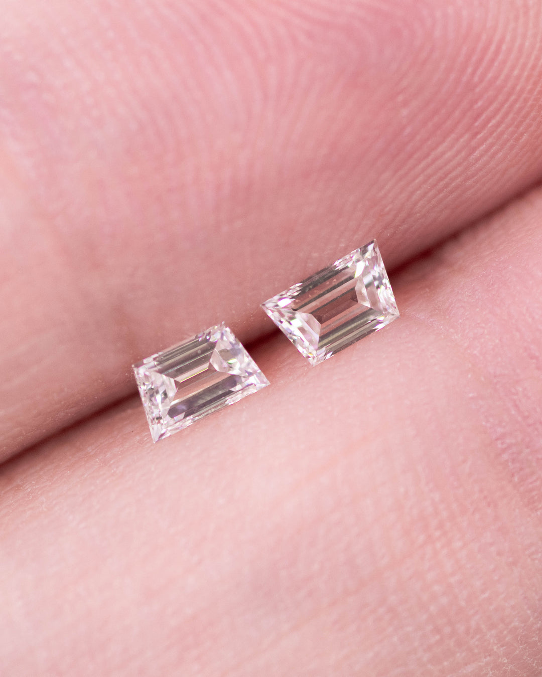 Close-up of 0.30 carat G color VVS clarity trapezoid step cut diamond pair from South Africa held on fingertips. The natural untreated diamonds showcase clean lines, crisp faceting, and perfect matching symmetry.