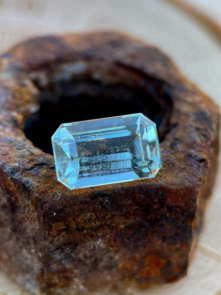 Emerald cut aquamarine gemstone with mint and blue hues placed on a rustic rock surface.
