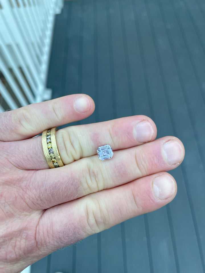 A 2.60-carat sapphire with a modified Asscher cut rests on a hand, displaying its vibrant purple and pink hues. The stone, measuring 7.71mm by 7.25mm by 4.97mm, originates from Sri Lanka and is untreated, no heat, reflecting its natural brilliance.