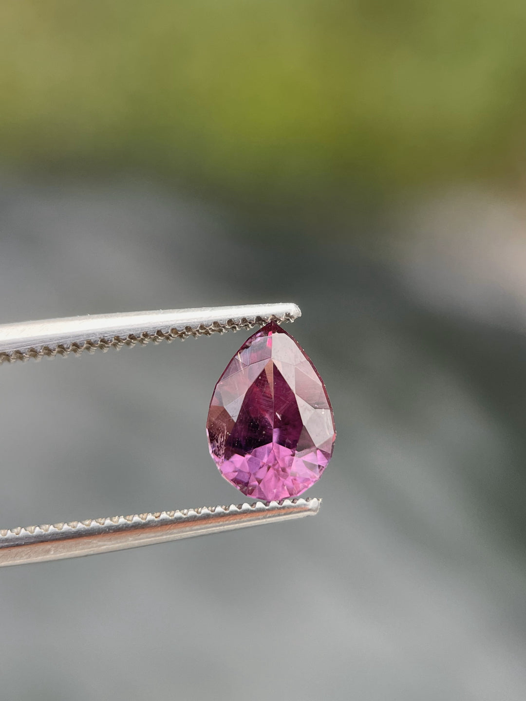 A pear-shaped pinkish-purple sapphire weighing 1.45 carats held by metal tweezers against a blurred green background.